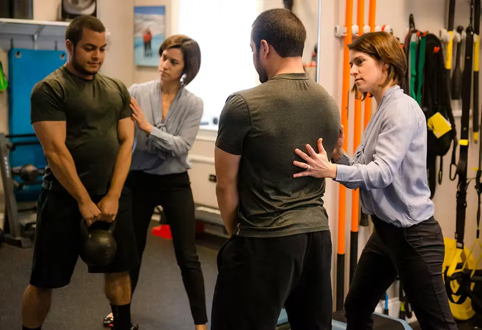 Owner Kristen Wilson with short brown hair, assisting a male patient lifting a kettle bell as a deadlift exercise
