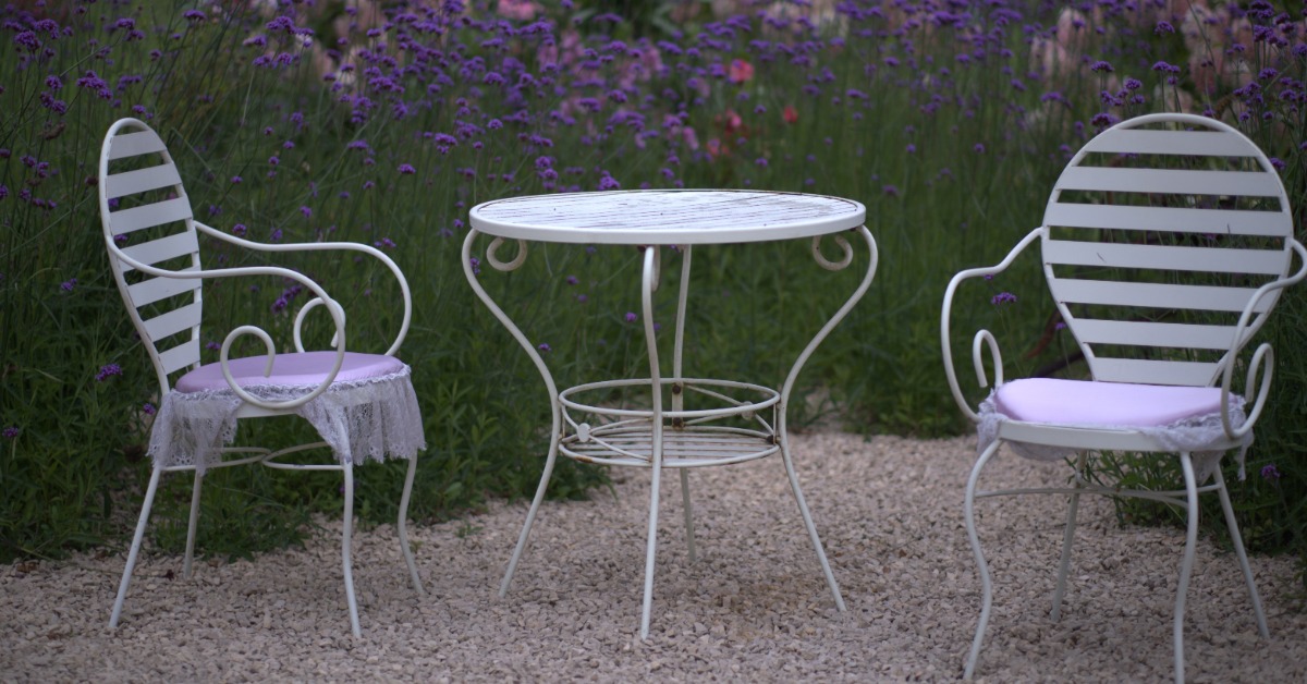 Two white metal patio chairs with lavender cushions flank a round white table on gravel, surrounded by purple flowers.