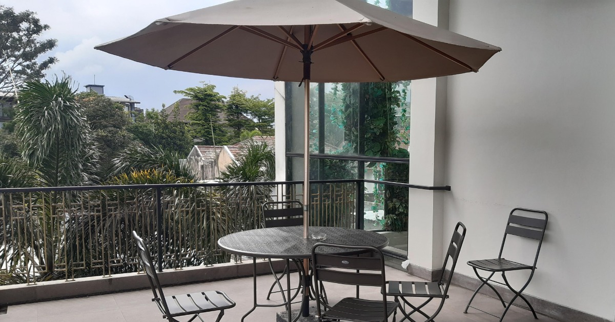 Black metal patio table and five folding chairs beneath a large brown umbrella on a balcony overlooking lush trees.