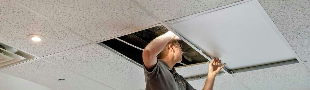 HVAC technician removing a ceiling tile to inspect ductwork and ventilation systems above a commercial drop ceiling.